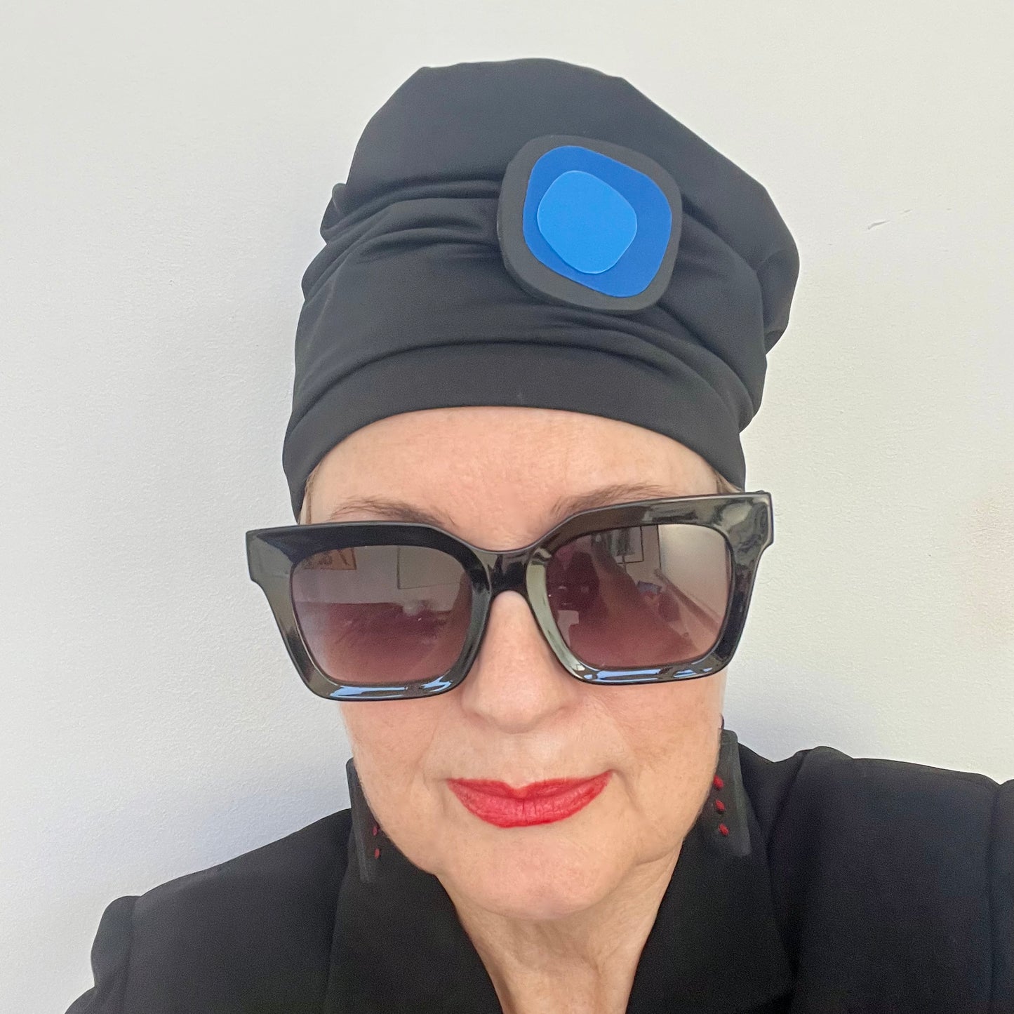 A woman wears a black hat topped with an irregular shaped black brooch base with 2 layers of blue shapes on top