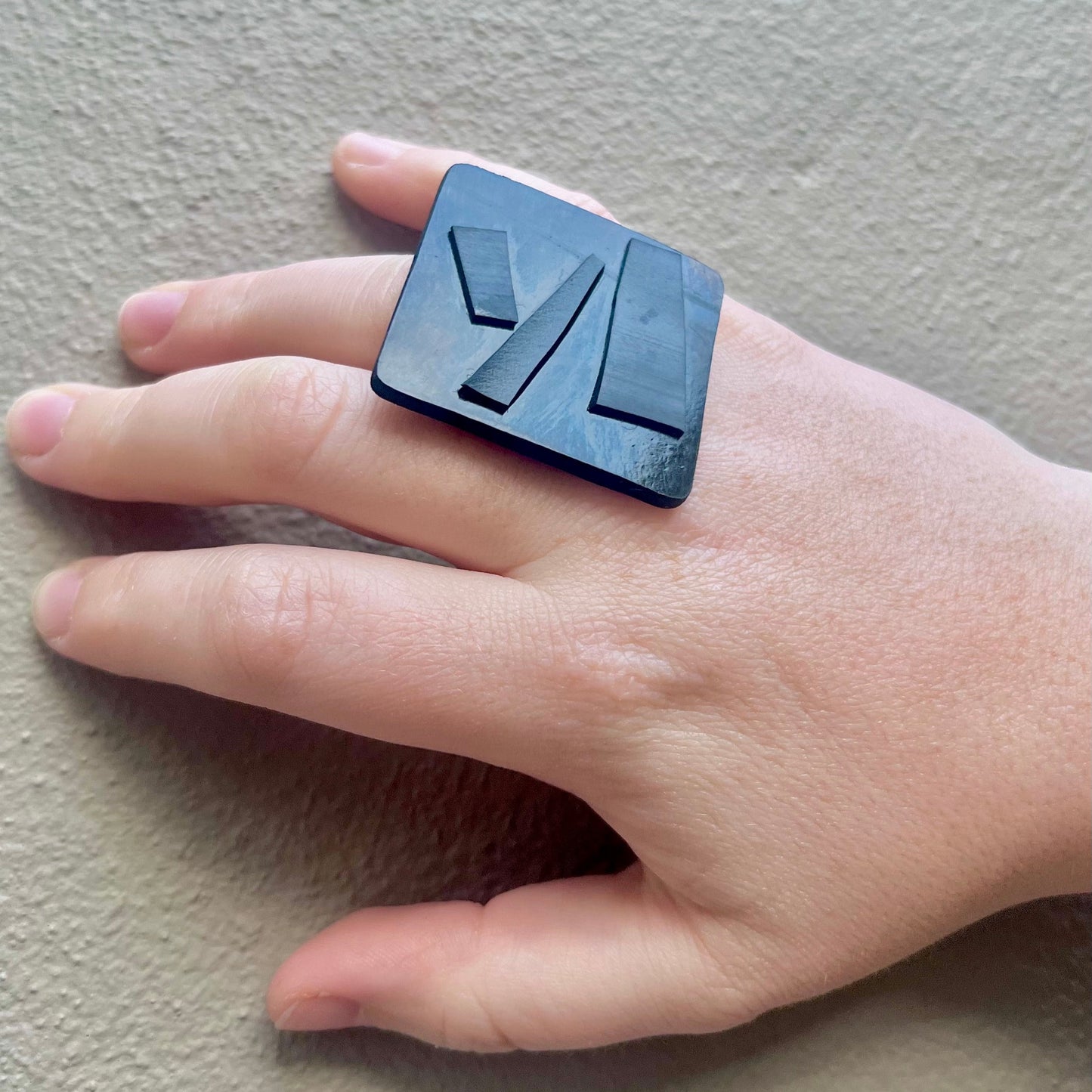 Hand wearing a black square ring with geometric design on a neutral background