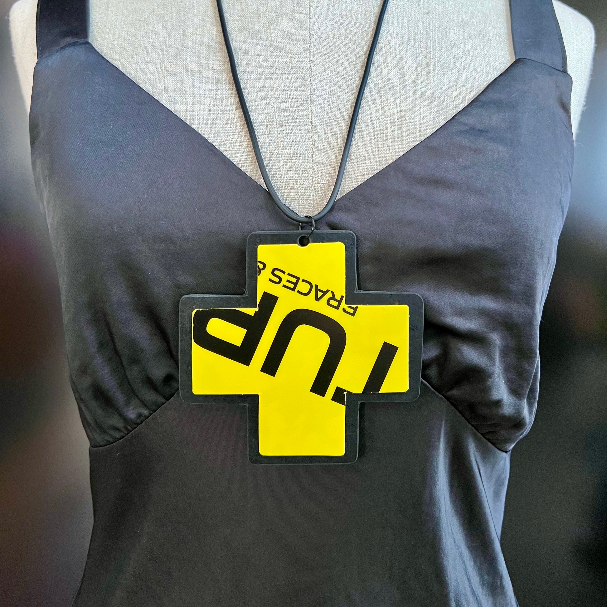 A pendant necklace on an angle is made from a black rubber disc topped with a yellow and black cross