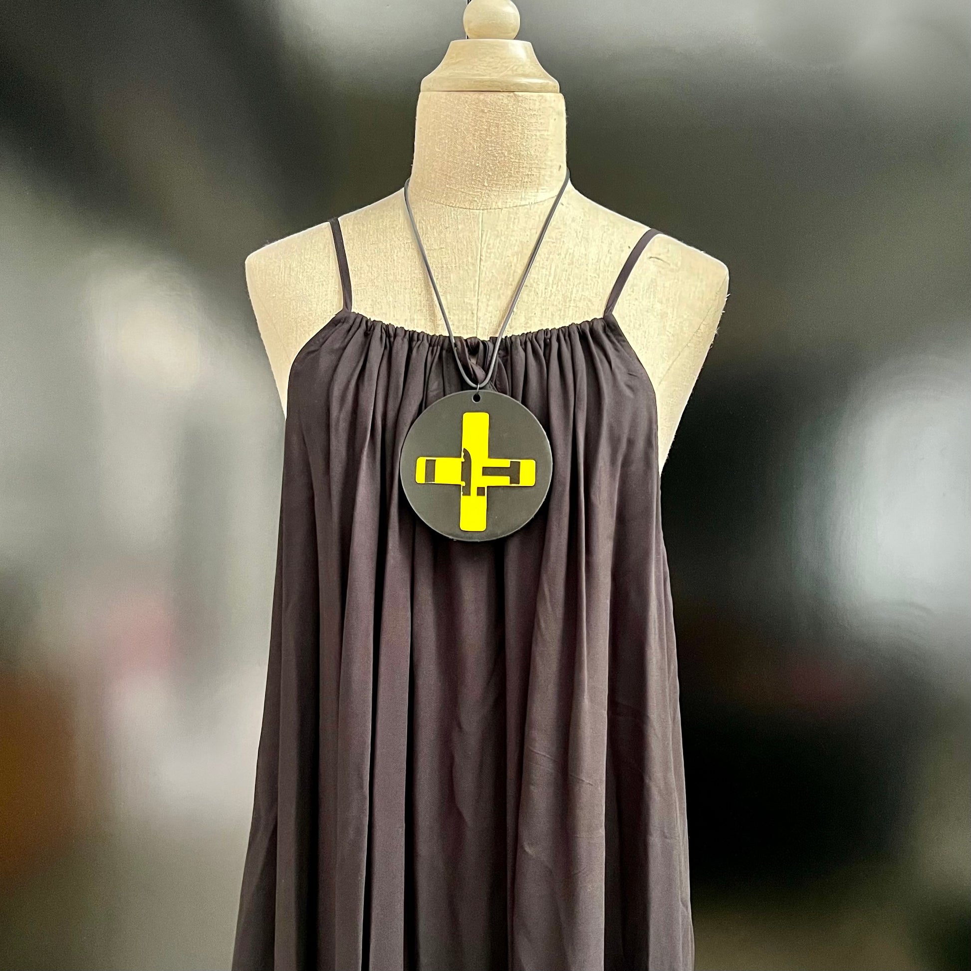 A pendant necklace on an angle is made from a black rubber disc topped with a yellow and black cross