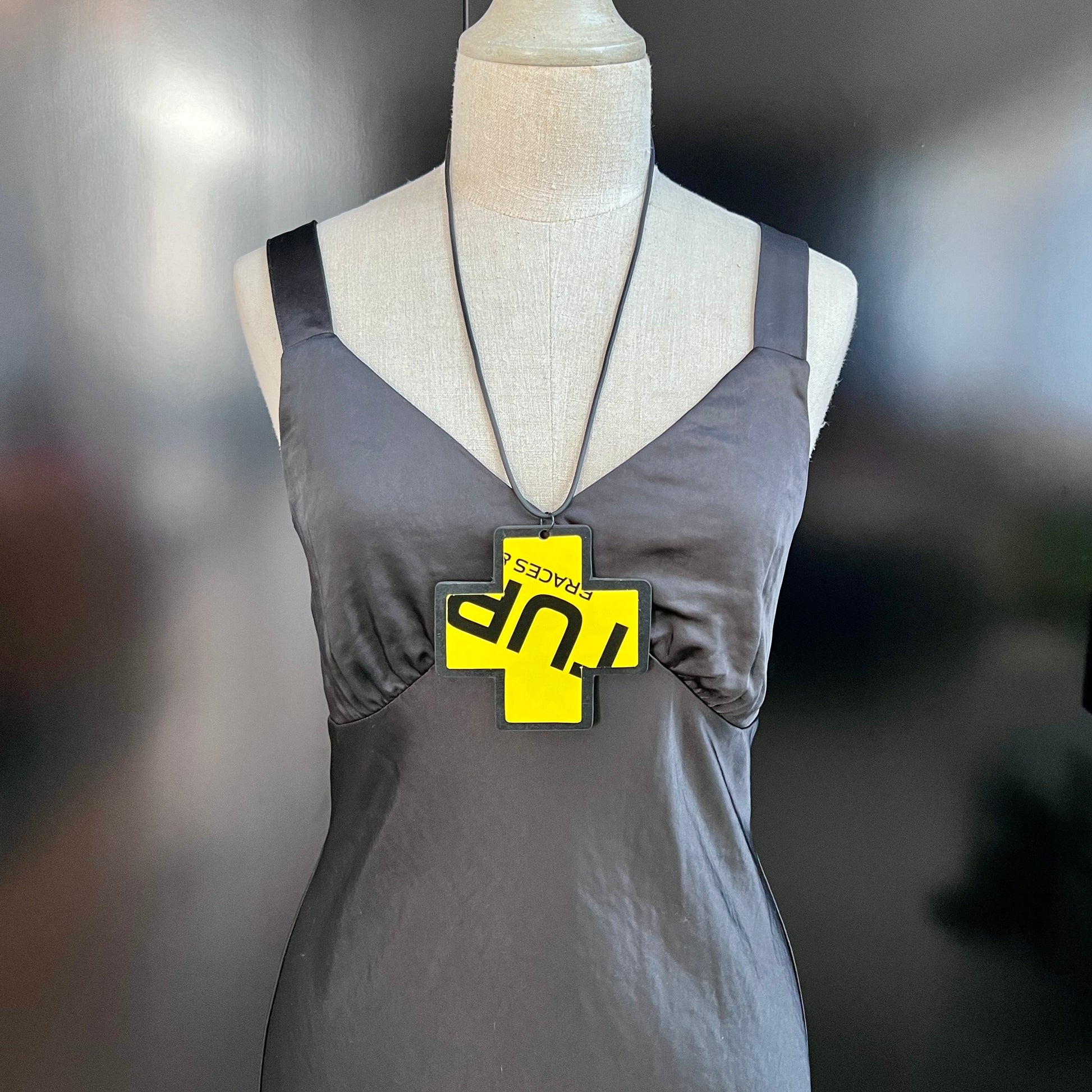 A mannequin wears a large pendant in the shape of a cross and layered with a black and yellow plastic cross with writing