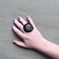 A hand wears a black ring topped with a bronze centre
