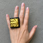 A hand wearing a large square ring with a crumpled gold center on a black base, set against a textured gray background.