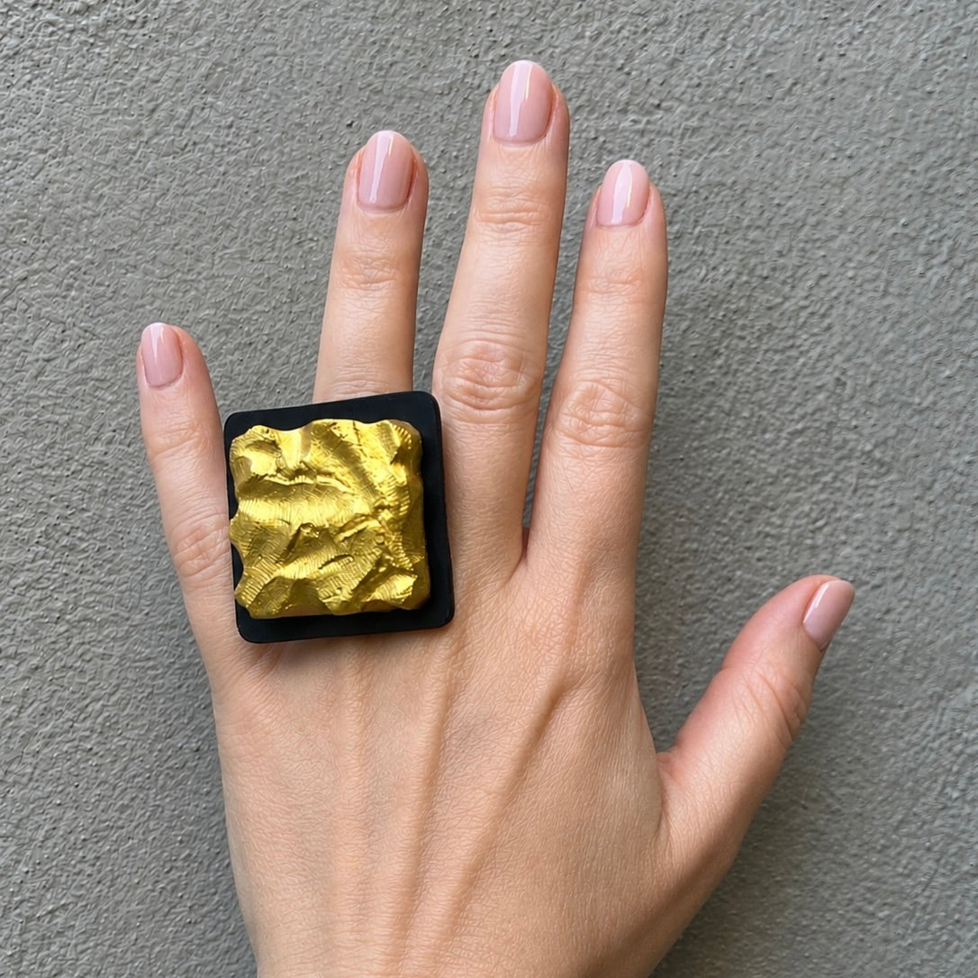 A hand wearing a large square ring with a crumpled gold center on a black base, set against a textured gray background.
