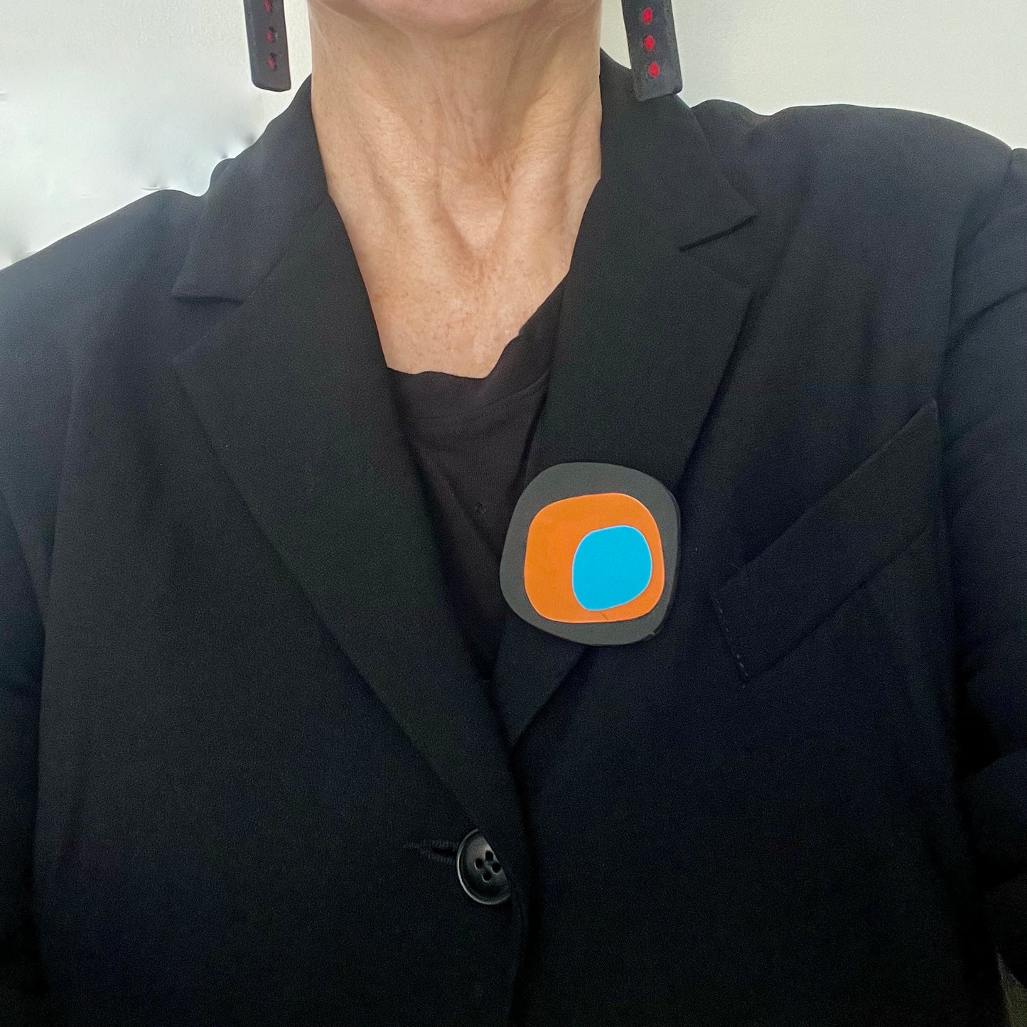 Person wearing a black blazer with a brooch with an irregular shaped black base topped with layers of orange and blue