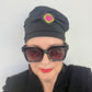 A woman wears a black hat topped with A black irregular shaped brooch layered with green and pink segments