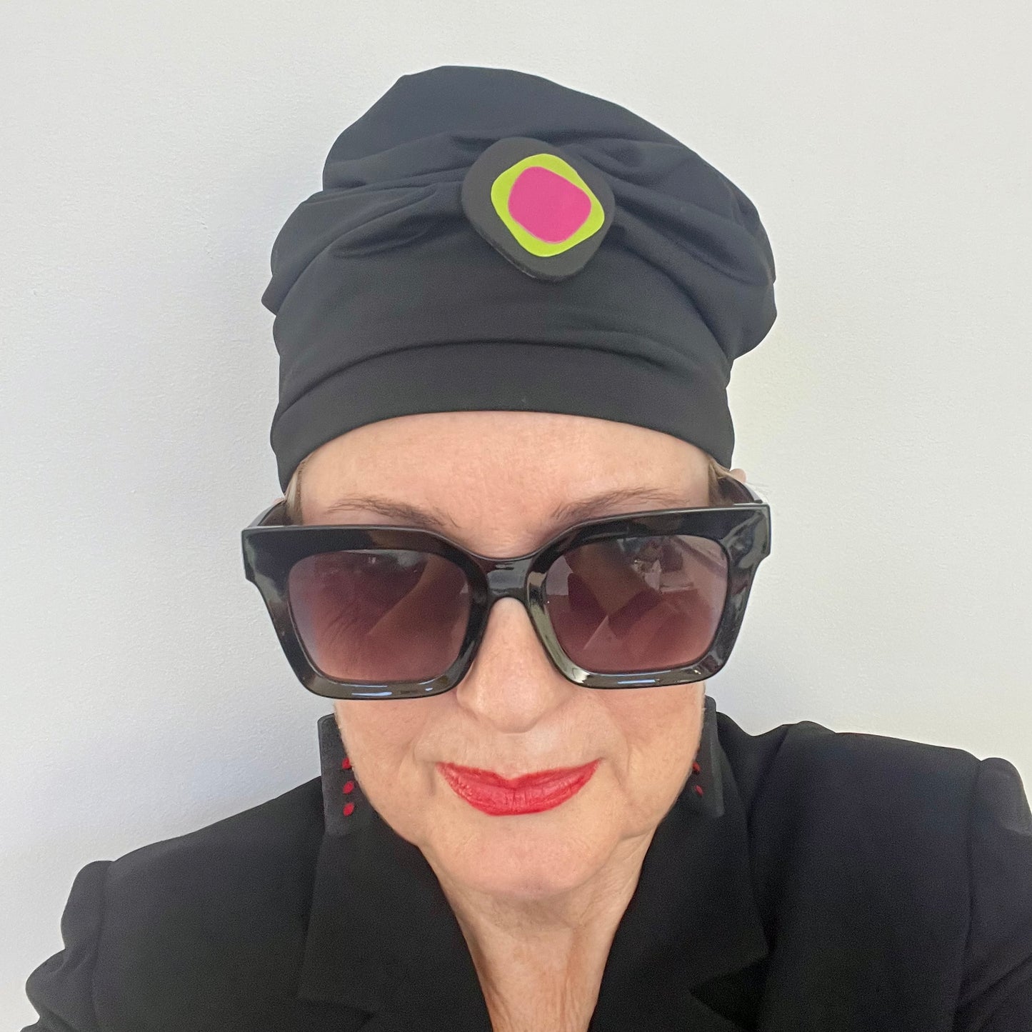 A woman wears a black hat topped with A black irregular shaped brooch layered with green and pink segments