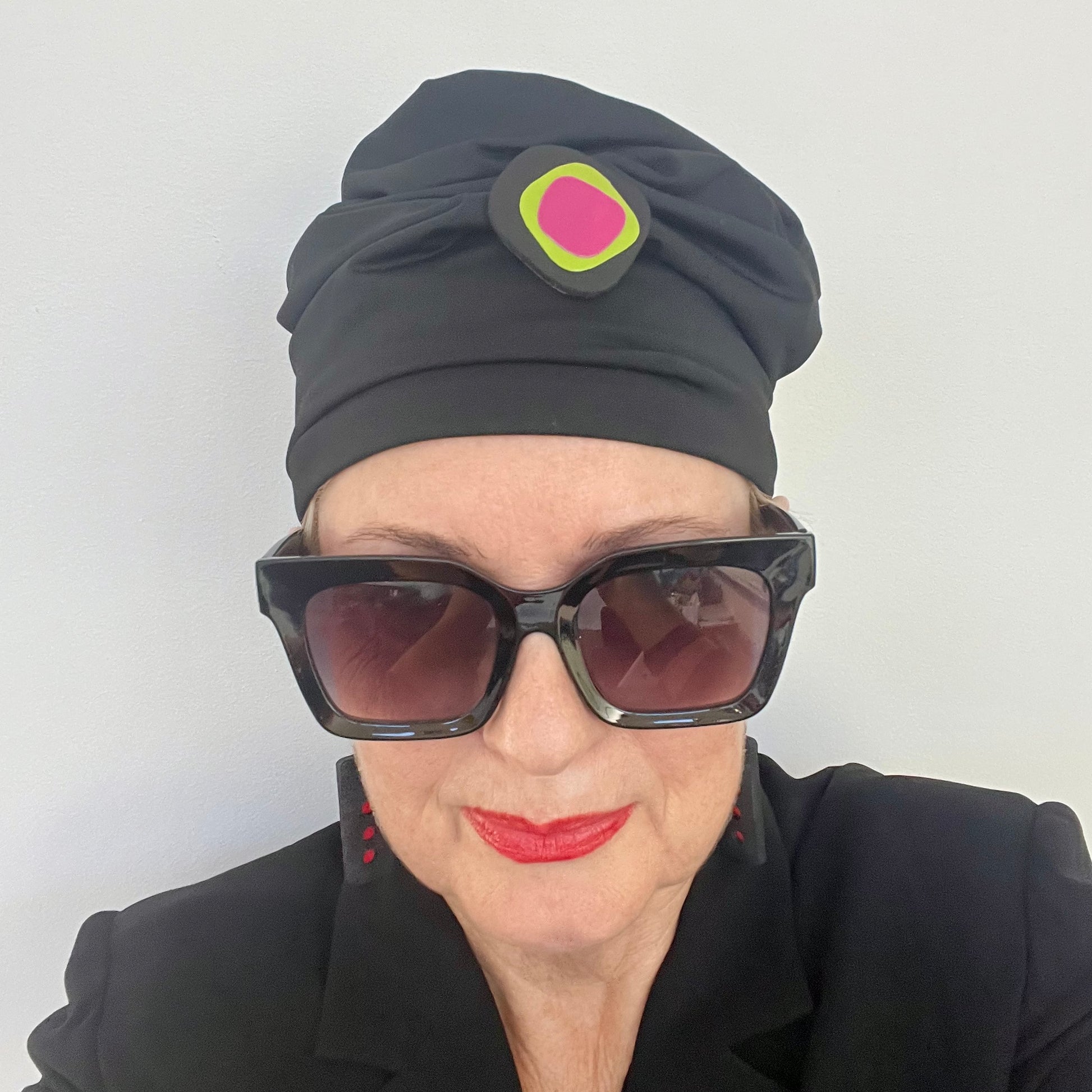 A woman wears a black hat topped with A black irregular shaped brooch layered with green and pink segments
