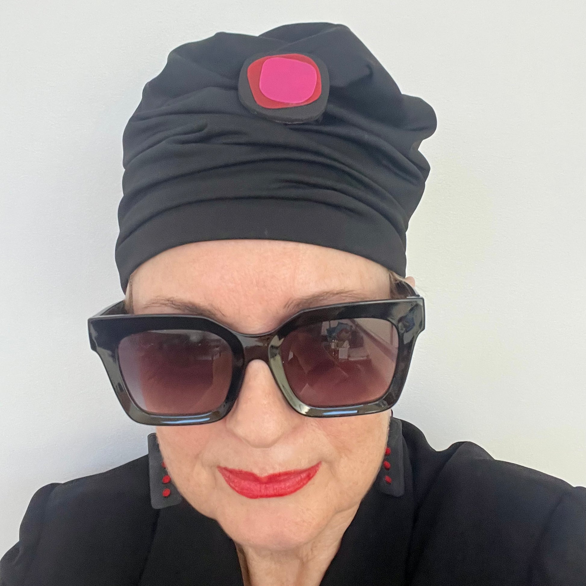 A woman wears a scrunchy black hat topped with A black irregular shaped brooch topped with layers of red and pink