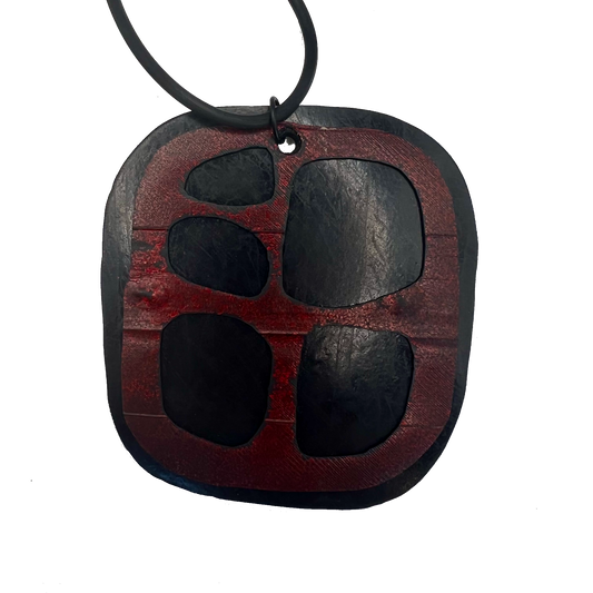 A misshapen square pendant made from black rubber is topped with a red rubber layer that has cutouts