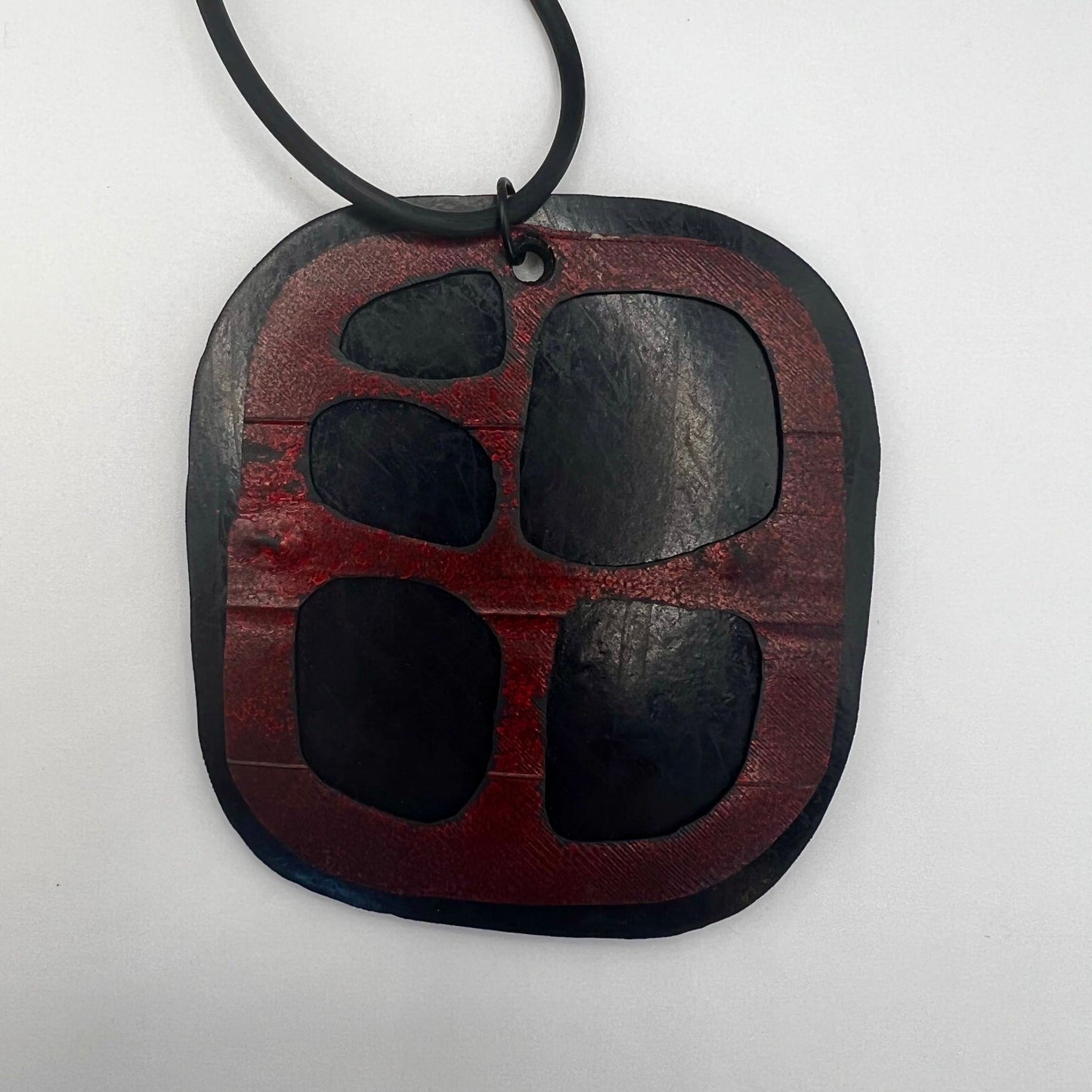 An organically shaped square pendant made from black rubber is topped with a red rubber layer that has cutouts.