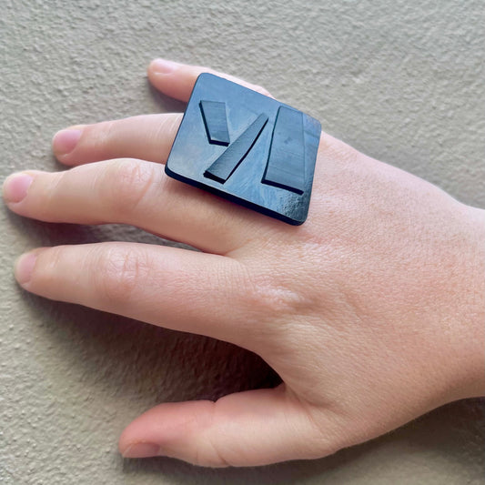 Hand wearing a black square ring with geometric design on a neutral background
