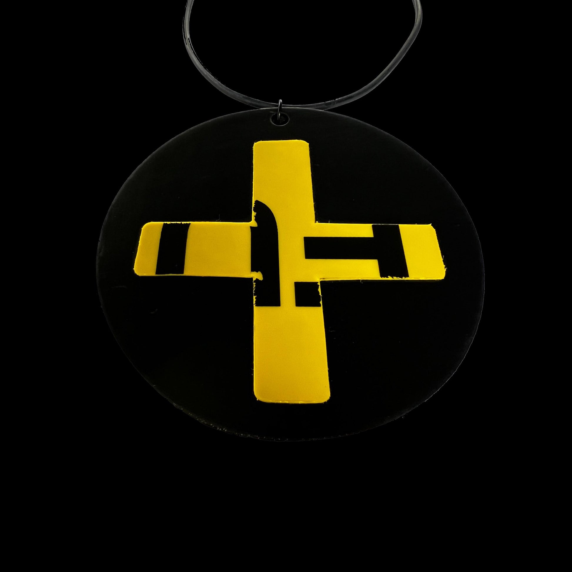 A pendant necklace on an angle is made from a black rubber disc topped with a yellow and black cross