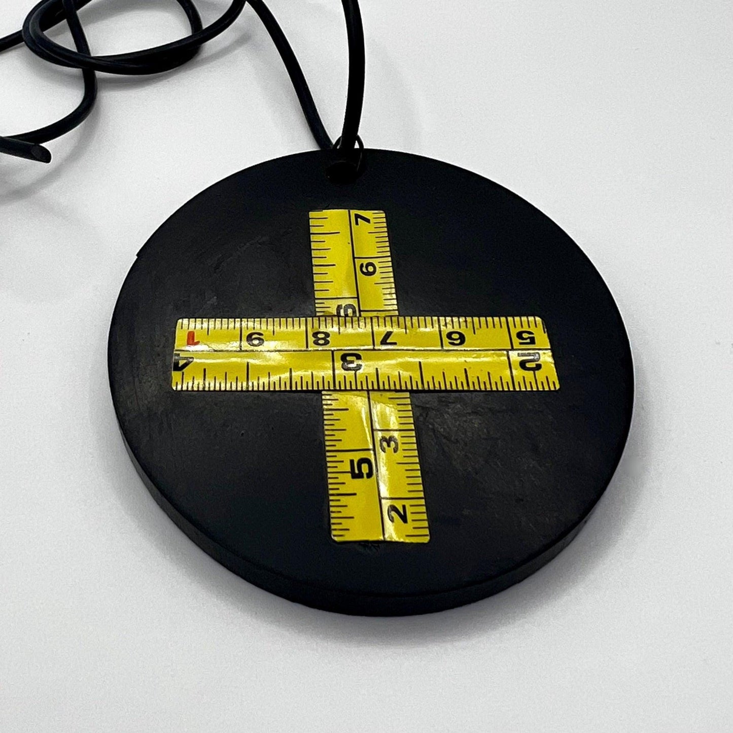 A round rubber black pendant with a yellow cross made from a metal tape measure