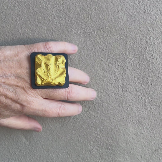 A hand wearing a textured gold square ring moves against a grey wall