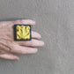 A hand wearing a textured gold square ring moves against a grey wall