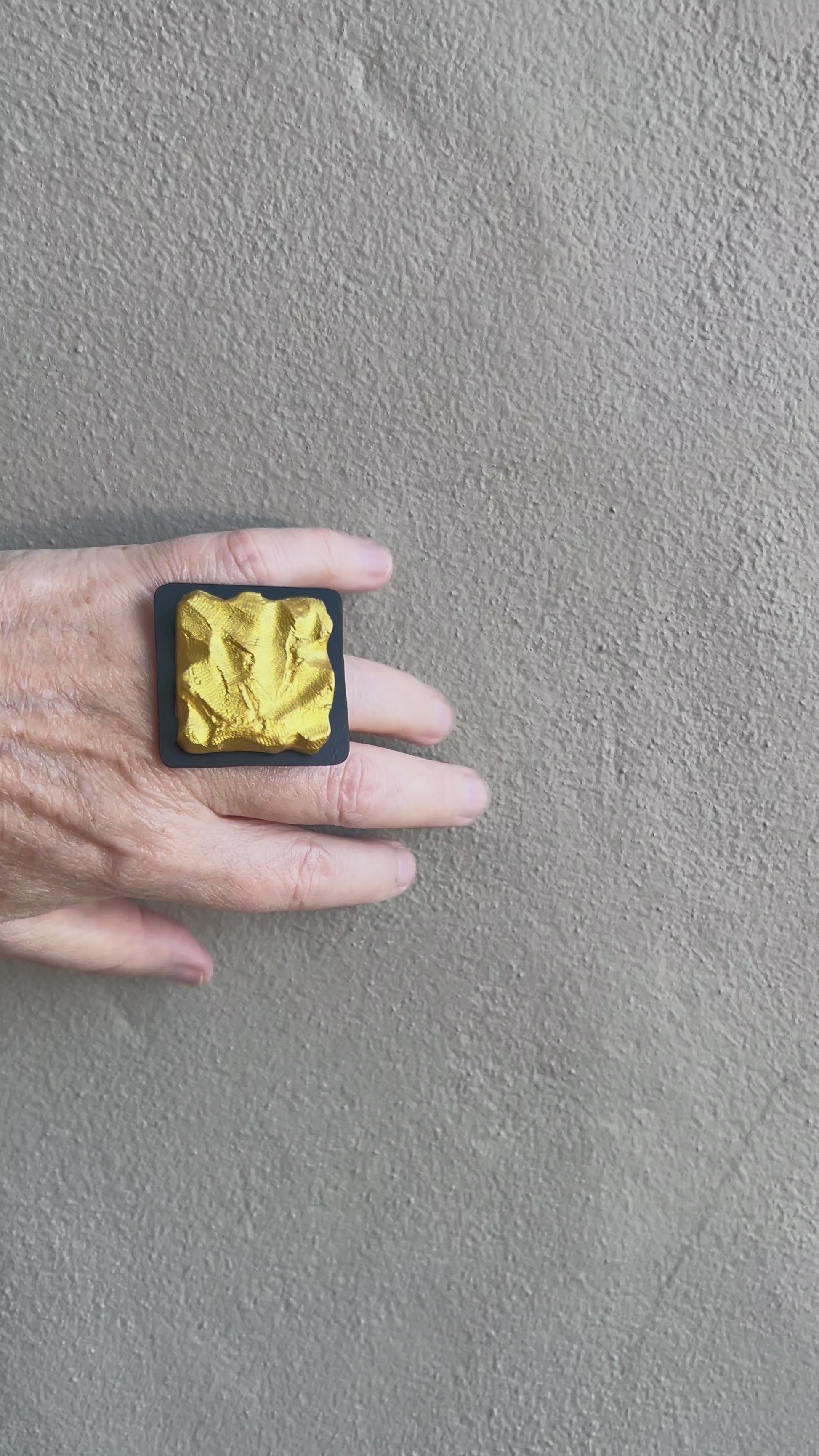 A hand wearing a textured gold square ring moves against a grey wall
