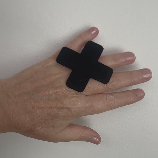 A hand wears a large cross shaped black ring